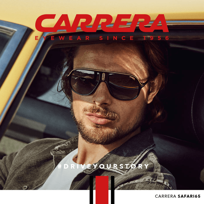 A man wearing black and gold Carrera Safari65 aviator sunglasses while sitting in a yellow vintage car, representing classic automotive eyewear trends.