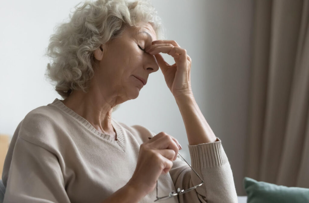 Elderly person rubs the bridge of her nose while holding eyeglasses, showing signs of eye strain or fatigue.