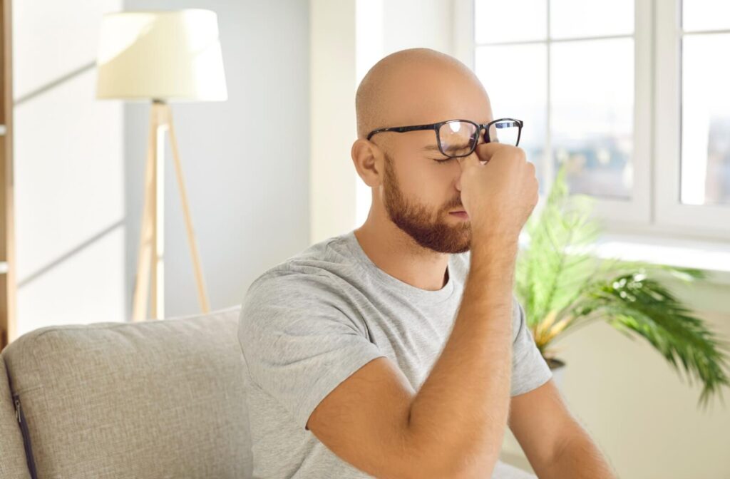 A person with a beard wearing glasses sitting on a sofa and rubbing the bridge of their nose with eyes closed, illustrating how dry eyes can cause vision blurriness and discomfort.