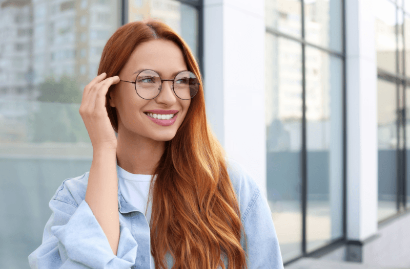 A smiling woman with long wavy red hair wearing round, clear prescription eyeglasses outdoors in natural light.