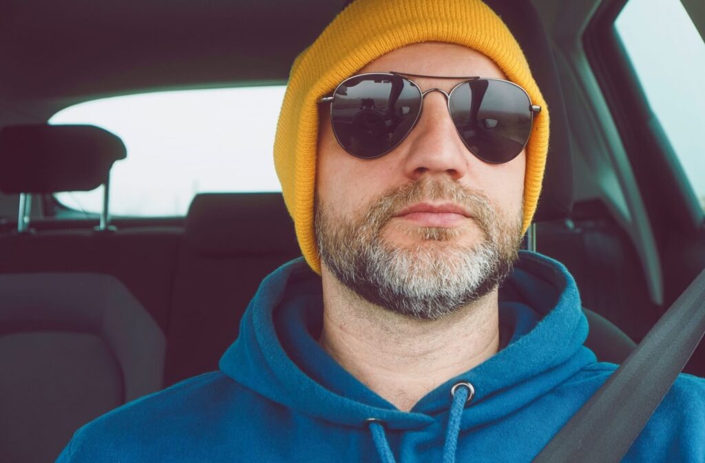 A man with a graying beard wearing a yellow beanie and dark aviator-style sunglasses while sitting in the driver's seat of a car.